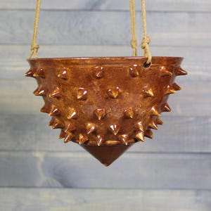 May include: A glossy brown ceramic hanging planter with a conical base and a textured surface covered in numerous sharp, protruding spikes. The bowl-shaped vessel is suspended by two tan rope cords attached to holes at the top rim.