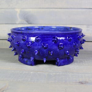 May include: A cobalt blue ceramic bowl with a textured surface and a series of conical spikes around the rim. The bowl has a low profile and sits on three small feet. The glaze has a glossy finish.