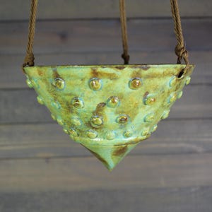 May include: A handmade, green ceramic hanging planter with a textured surface of small, raised dots. The planter has a conical shape and is suspended by three brown rope cords. The glaze has brown and blue accents.