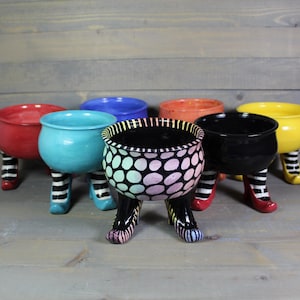 May include: Six colorful ceramic pots with legs shaped like witch's shoes. The pots are decorated with polka dots and stripes.