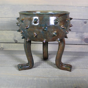May include: A handcrafted ceramic planter with a unique design. The brown and teal-colored pot features a textured surface with protruding spikes and is supported by three leg-like structures. The planter is suitable for indoor or outdoor use.