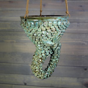 May include: A hanging ceramic planter featuring a textured, scale-like surface in shades of teal, blue, and green. The vessel has a wide, circular opening at the top and a curved, tail-like extension at the bottom, suspended by three tan ropes.