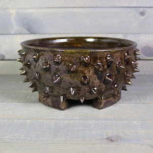 May include: A handcrafted, brown ceramic bowl with a textured surface and numerous, evenly spaced, metallic spikes. The bowl has a wide opening and sits on three small, square feet. The interior has a glossy finish.