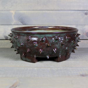 May include: A handcrafted ceramic bowl with a unique design. The bowl is a deep burgundy color with a teal glaze and is adorned with pointed, spike-like protrusions around the exterior. The bowl has a low profile and sits on three small feet.