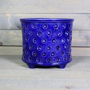 May include: A cylindrical ceramic planter in a deep cobalt blue glaze, featuring a repeating pattern of small, circular indentations across the exterior surface. The vessel is supported by three short, rounded feet at the base.