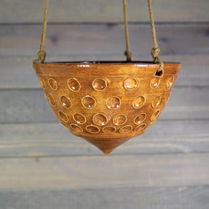 May include: A hanging planter made of brown ceramic with a series of circular indentations. The planter is suspended by a natural rope. The planter has a conical bottom and a wide opening, suitable for plants.
