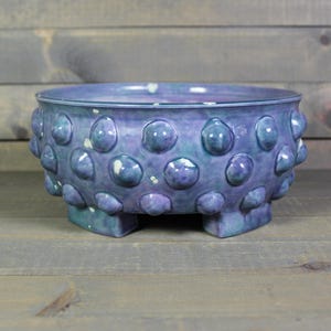 May include: A ceramic bowl with a textured surface and a unique design. The bowl is a blend of blue and purple hues, with a series of rounded, raised bumps around the exterior. It rests on three small, square feet.