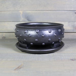 May include: A black ceramic planter with a matching saucer. The planter has a wide, shallow bowl shape with a series of raised, rounded bumps around the rim. The saucer has a slightly raised edge and three small feet. The set is made of a dark, matte ceramic.