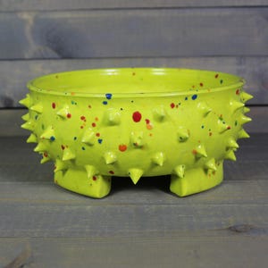 May include: A chartreuse ceramic bowl with a textured surface and a series of conical spikes around the rim. The bowl is speckled with red, blue, and orange paint. The bowl is supported by three square feet.