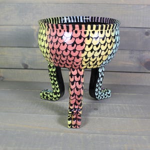May include: A colorful ceramic bowl with a unique design. The bowl features a rainbow-colored scale pattern on the exterior and is supported by three black legs. The rim is black with vertical stripes. The bowl is a decorative item.
