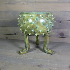 May include: A ceramic planter with a rounded body covered in numerous small, sharp spikes. The vessel features a mottled green and brown glaze and is supported by three long, curved legs that extend outward from the base of the bowl.
