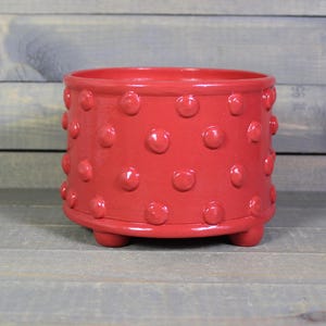 May include: A vibrant red ceramic planter with a textured, dotted pattern. The planter has a rounded shape, sits on three small feet, and is suitable for indoor or outdoor use. The planter is approximately 6 inches in diameter and 5 inches tall.
