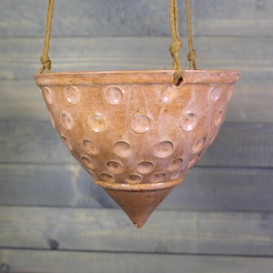 May include: A conical ceramic hanging planter with a light brown, glazed finish. The exterior surface features a repeating pattern of circular indentations. The planter is suspended by three thick, tan-colored twisted rope cords attached to the rim.