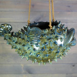 May include: A ceramic pufferfish-shaped hanging planter with a glossy blue and green glaze. The fish features numerous pointed spikes across its body, a defined tail fin, and a side fin. Two tan ropes are attached to the top for hanging purposes.