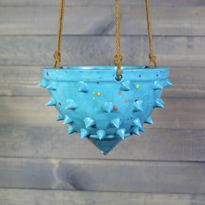 May include: A turquoise ceramic hanging planter with a textured, spiky exterior. The planter has a speckled design with orange, yellow, and red accents. It is suspended by three brown rope strands, ideal for displaying plants.