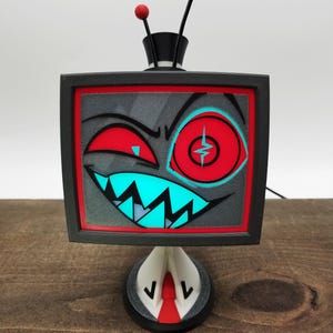Puede incluir: Una figura de personaje de televisión impresa en 3D con marco negro, una pantalla que muestra un gráfico rojo y cian de una cara guiñando un ojo con dientes afilados, y dos antenas negras con esferas rojas en la parte superior sobre una base blanca y roja.