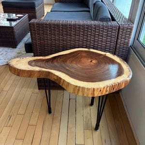 May include: A live edge wood slab side table featuring a polished, dark brown circular grain pattern with a lighter natural wood border. The tabletop is supported by four thin, black metal hairpin legs that extend outward from the center.