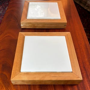 May include: Two square wooden frames with light brown grain patterns, each containing a smooth, flat, white square tile insert. The frames feature mitered corners and a clean, simple construction, presenting a neutral and functional design.