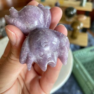 May include: A carved, lavender-colored crystal crab figurine. The crab has a detailed, textured surface and is held in a hand. The figurine is a unique decorative item, possibly for a crystal collection or home decor.