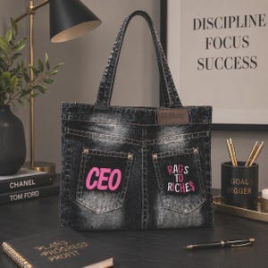 May include: Black denim tote bag with a design resembling jean pockets. The bag features the word "CEO" in pink on one pocket and "Rags to Riches" on the other. The bag has a top handle and a "Shifang" label.
