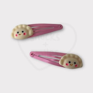 May include: Two pink plastic hair clips with a white smiling dumpling design.