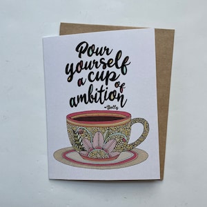 May include: A white greeting card featuring a colorful, intricately patterned teacup on a saucer. The text reads, "Pour yourself a cup of ambition ~Dolly" in black script lettering. The cup displays pink, yellow, and brown floral designs.