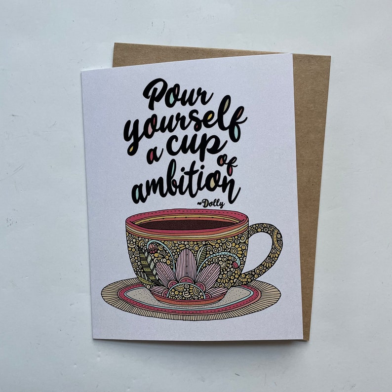 May include: A white greeting card featuring a colorful, intricately patterned teacup on a saucer. The text reads, "Pour yourself a cup of ambition ~Dolly" in black script lettering. The cup displays pink, yellow, and brown floral designs.