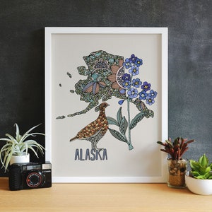 May include: A white framed print with a stylized illustration of the state of Alaska. The illustration is filled with intricate patterns and colors. A brown and white bird is perched on a branch with blue flowers.