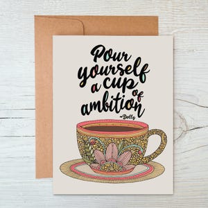 May include: A greeting card with a light beige background featuring a black script that reads Pour yourself a cup of ambition ~Dolly above a detailed illustration of a teacup and saucer decorated with intricate floral and geometric patterns.