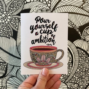 May include: A white greeting card featuring a black, yellow, and pink illustration of a teacup on a saucer with intricate floral patterns. The text reads, "Pour yourself a cup of ambition" followed by "~Dolly" in a black script font.