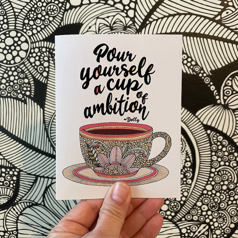 May include: A white greeting card featuring a black, yellow, and pink illustration of a teacup on a saucer with intricate floral patterns. The text reads, "Pour yourself a cup of ambition" followed by "~Dolly" in a black script font.