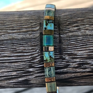 May include: A beaded bracelet with a mix of square and rectangular beads in shades of teal, gold, and bronze. The bracelet is displayed against a weathered wooden surface, showcasing its colors and textures.
