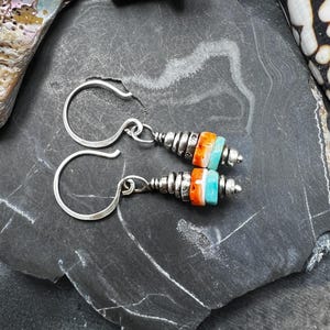 Op de afbeelding: Een paar zilveren oorhangers met een stapel kleine, cilindrische kralen in levendig oranje, wit en turquoise. De oorbellen zijn voorzien van zilverkleurige tussenstukjes en hebben een eenvoudig ontwerp met een gebogen zilveren haak.