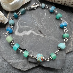 Op de afbeelding: Handgemaakte armband met kleurrijke glazen kralen in blauw- en groentinten, verbonden door een zilverkleurige ketting. De armband is ongeveer 18 cm lang. De kralen variëren in vorm en grootte.