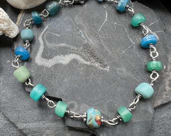 May include: A handmade bracelet featuring a variety of colorful glass beads in shades of blue and green, strung together with a silver-colored chain. The bracelet is approximately 7 inches long.