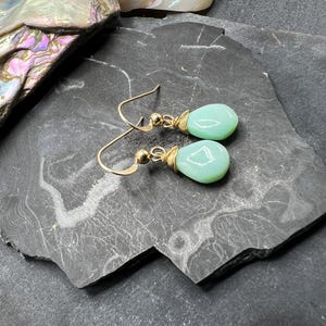 May include: A pair of drop earrings featuring smooth, teardrop-shaped light green stones. Each stone is wrapped in thin gold-toned wire at the top and attached to a gold-toned hook ear wire with a small bead accent near the connection point.