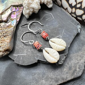 May include: Silver hook earrings featuring white seashell pendants and coral-colored beads. The earrings are set against a dark gray stone surface, with additional seashells and coral in the background. The earrings are a beach-themed accessory.