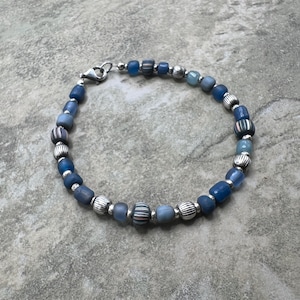 May include: A beaded bracelet featuring a mix of blue glass beads and silver-colored metal beads. The bracelet has a variety of blue hues, with some beads striped in white, red, and blue. The bracelet is secured with a silver-colored clasp.