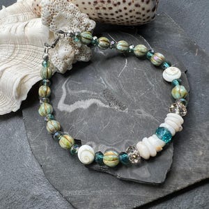 May include: A handmade bracelet featuring a mix of natural elements. The bracelet includes green and gold-toned beads, clear crystal beads, and white shell accents. The bracelet is secured with a silver-toned clasp.