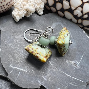 May include: Handmade earrings with speckled, light green and brown glass beads. The earrings have a silver hook and small, round, light green beads. The earrings are displayed on a dark grey, textured surface. The earrings are a unique accessory.