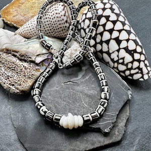 May include: A black and white beaded necklace with a silver clasp. The necklace features rectangular black beads with white stripes, and a section of white, irregular beads. The necklace is displayed on a dark gray surface with seashells.