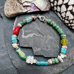 Peut inclure: Un bracelet composé de perles multicolores de formes et matériaux variés, incluant turquoise, corail rouge, coquillage blanc, verre vert et une perle verte en forme de poisson avec des détails dorés, fermé par un fermoir mousqueton argenté.
