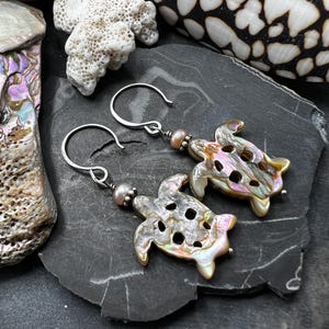 May include: A pair of silver earrings featuring iridescent, shell-shaped turtle pendants. Each earring has a small, round, pinkish bead and a silver hook. The earrings are displayed on a dark, textured surface, with seashells in the background.