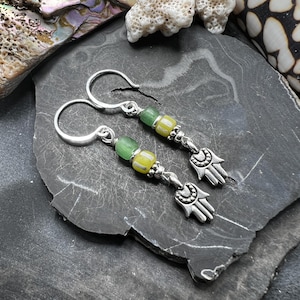 May include: Silver drop earrings featuring green and yellow beads and a Hamsa hand charm. The earrings have a hook fastening and are shown on a dark grey, textured surface. The beads are a mix of opaque and translucent glass.