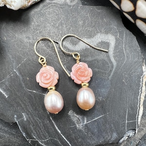 May include: A pair of gold-colored drop earrings. Each earring features a light pink rose-shaped stone above a pear-shaped, light pink pearl. The earrings are displayed on a dark gray, textured surface.