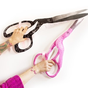 May include: Two large decorative scissors. One is black with a floral pattern, the other is pink with a bird-like design. Both have silver blades and are held against a white background.