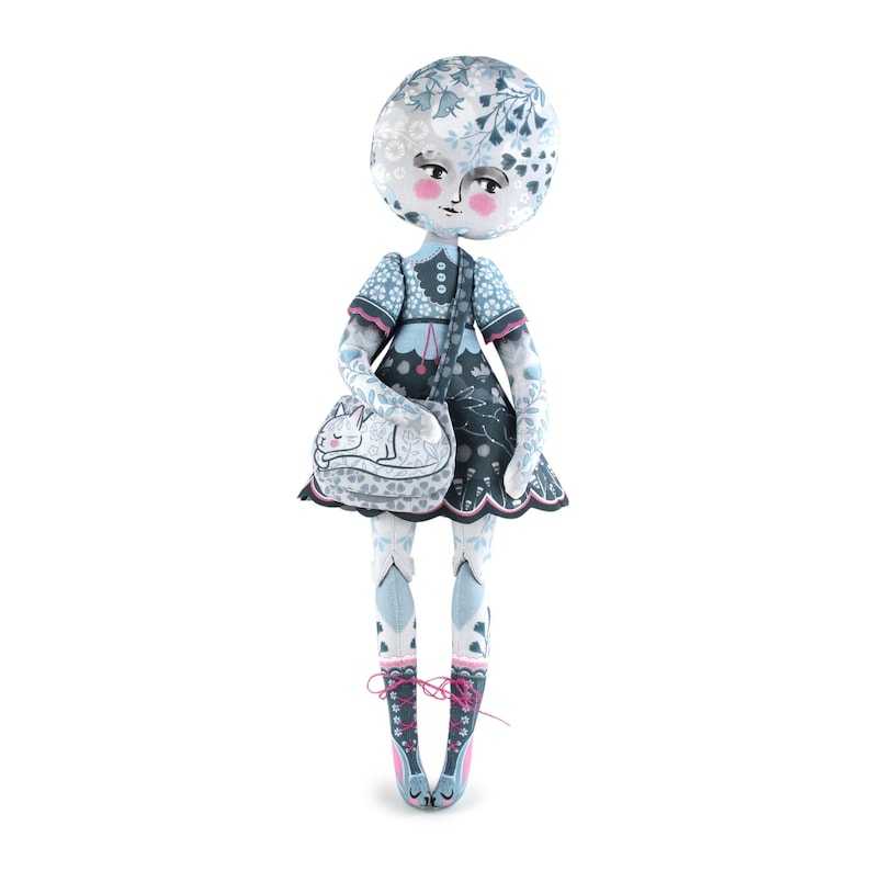 K&ouml;nnte beinhalten: Eine skurrile Puppe mit rundem Kopf und floralem Muster. Die Puppe tr&auml;gt ein blaues Kleid, eine passende Tasche mit Katzenmuster und Ballettschuhe. Das Gesicht der Puppe hat rosige Wangen und einen neutralen Ausdruck.