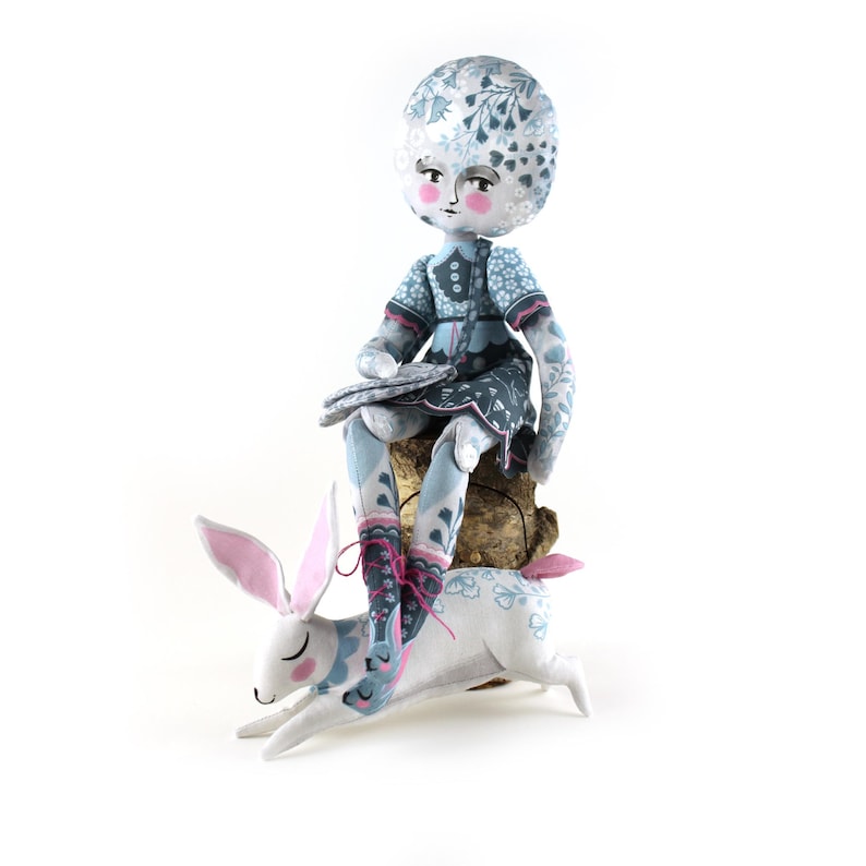 K&ouml;nnte beinhalten: Eine handgefertigte Puppe mit floral gemustertem Kopf und Kleid, die auf einem Holzsockel sitzt, mit einem wei&szlig;en Kaninchen darunter. Die Stiefel der Puppe und der K&ouml;rper des Kaninchens weisen ein &auml;hnliches Blumenmuster auf. Das Kaninchen hat rosa Ohren und einen rosa Schwanz.