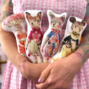 May include: Four flat fabric dolls featuring printed illustrations of a mushroom-headed figure, a fox, a cat, and a mouse. Each doll wears detailed, colourful clothing patterns including florals and hearts, with soft, rounded shapes and distinct facial features.