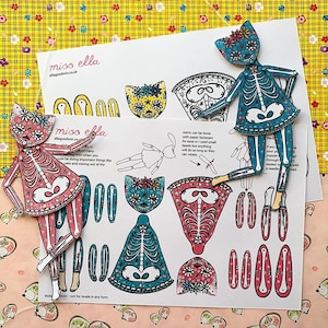 May include: Two colorful paper cat skeleton puppets with floral designs, alongside a sheet of cut-out pieces and instructions. The puppets are pink and blue, with intricate patterns and a whimsical aesthetic. The background features a yellow floral pattern.